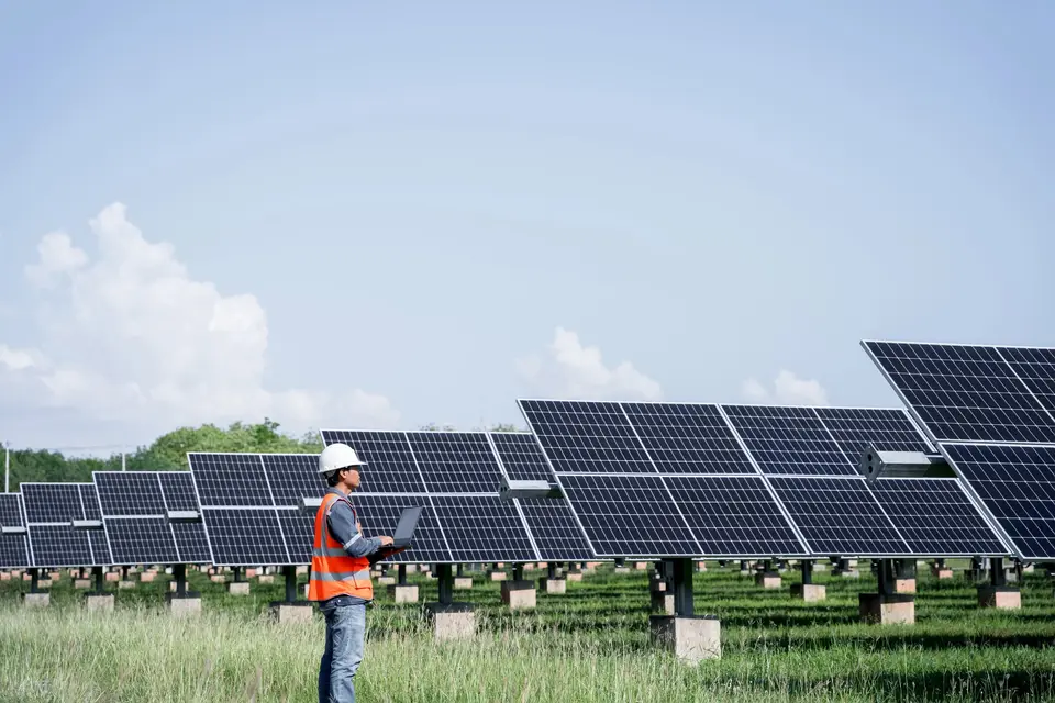 Technician in a hard hat checking plant data on a laptop at a ground-mounted array